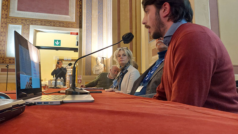 Presentazione pubblica: relatore al tavolo con microfono e slide