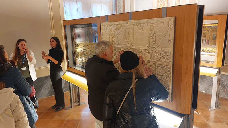 Fruizione tattile: visitatori esplorano una tavola in rilievo davanti alla teca